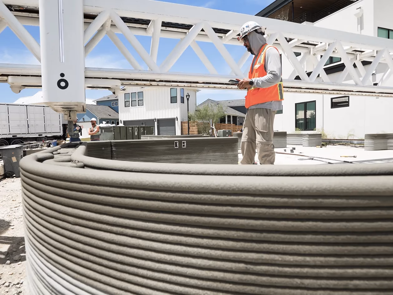 Worker in orange vest beside gantry 3D printer and layered concrete wall