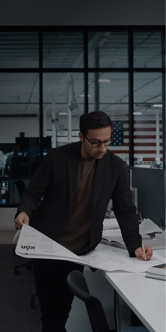 Man in glasses reviewing architectural plans at an office desk with workshop behind glass.