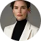 Portrait of a young woman with short brown hair wearing a black turtleneck and light beige blazer against a gray background.