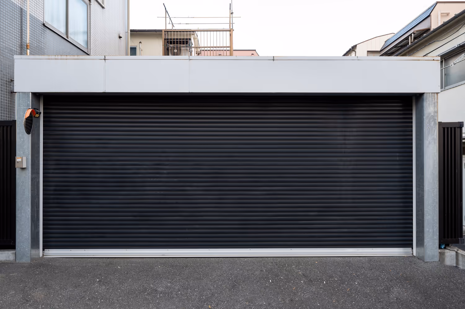 Closed black rolling garage door in a residential area with grey walls and pavement.
