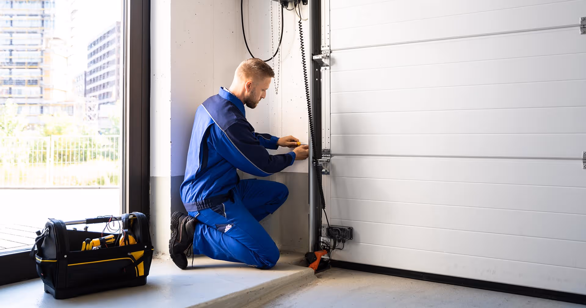 Technician in blue uniform kneeling and repairing the mechanism of a white garage door next to a toolbox.