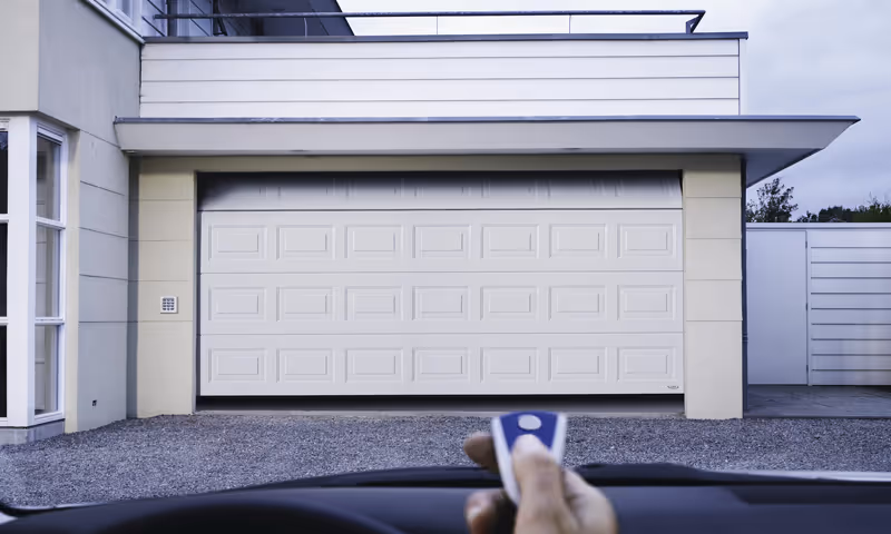 View from inside a car of a white automatic garage door with a hand holding a remote control in the foreground.