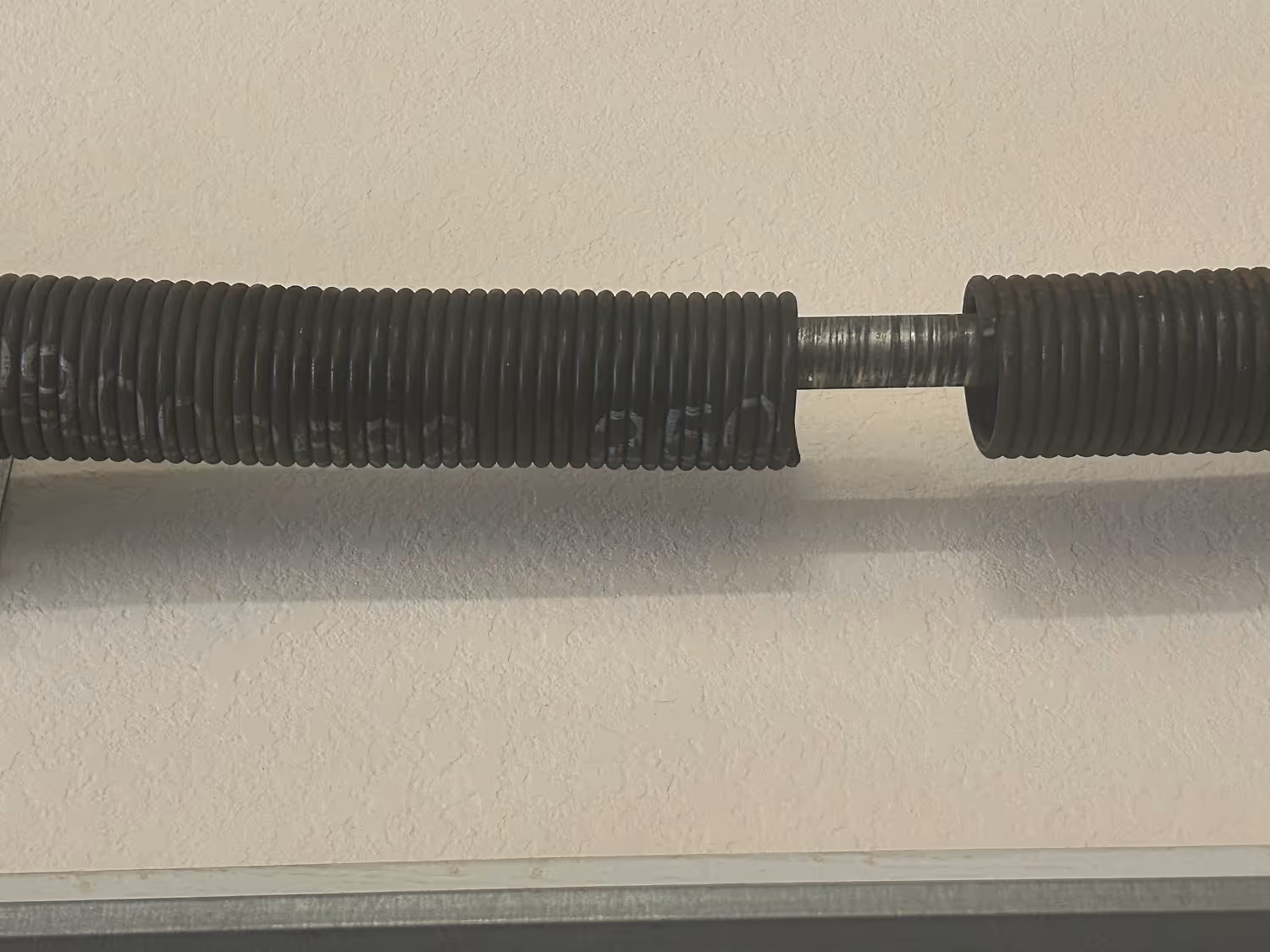 Close-up of a broken garage door torsion spring mounted on a metal rod against a textured beige wall.