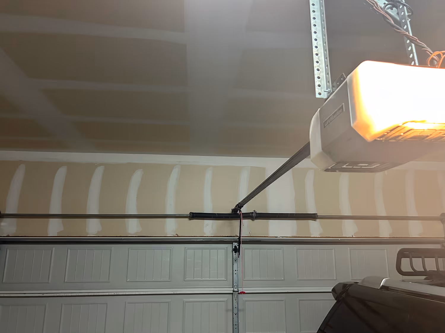 Interior of a garage showing a closed white garage door, an automatic garage door opener, and unfinished drywall.