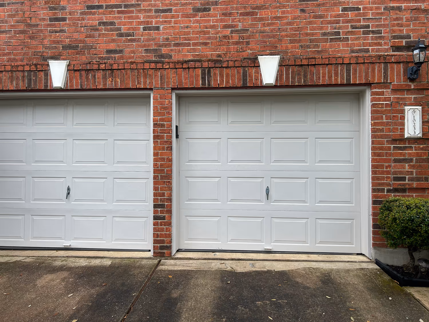 Two white garage doors set in a red brick wall with a driveway in front and a small bush on the right side.