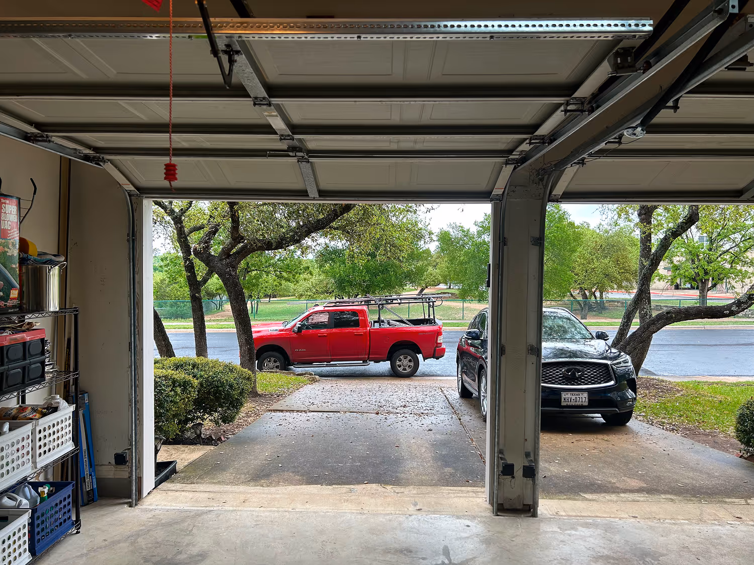 View from inside a garage looking out onto a driveway with a red pickup truck and a black SUV parked outside, with trees and greenery in the background.