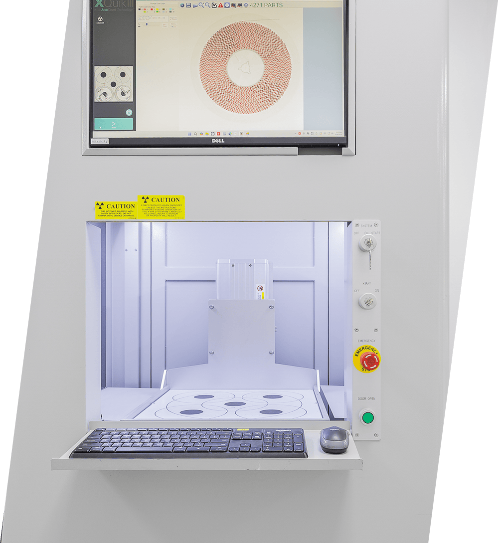 Equipamento de inspeção com monitor Dell mostrando um círculo composto por várias partes em vermelho, teclado e rato, botões de controlo e avisos de cautela para radiação.