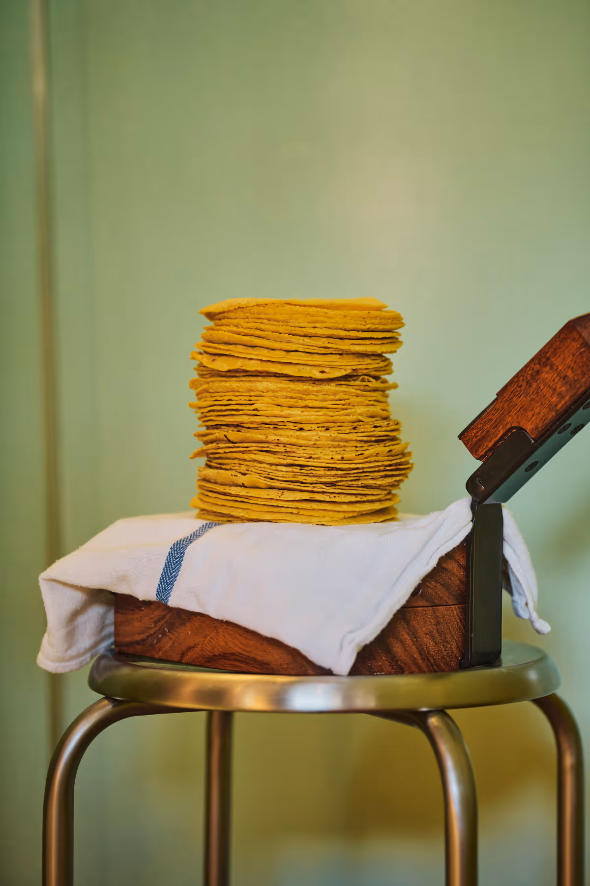 Stack of yellow corn tortillas on a white cloth inside a wooden and metal container on a stool.