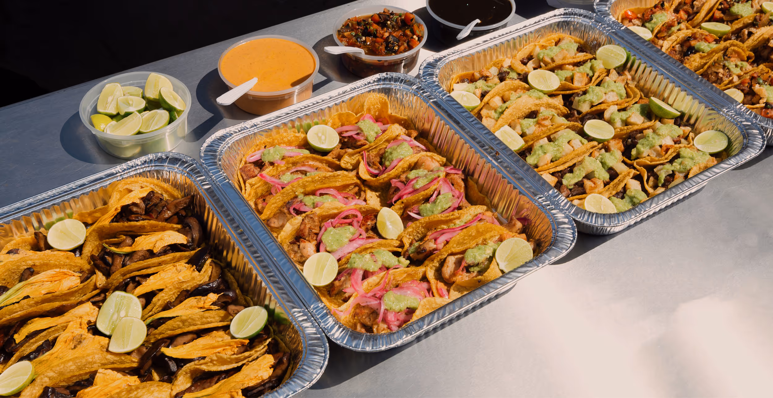 Trays of assorted tacos with lime wedges, served with containers of sauce and salsa on a metal table.