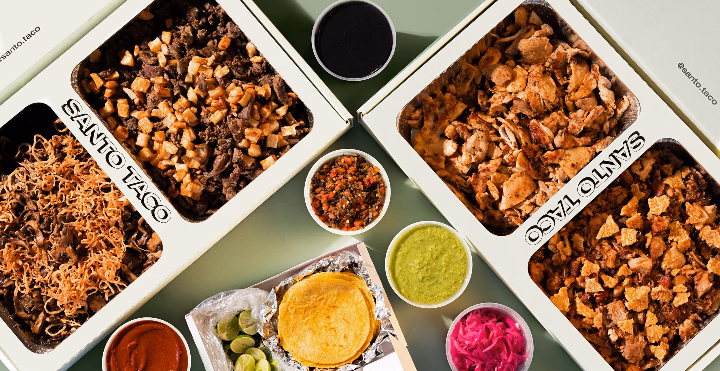 Trays of assorted tacos with lime wedges, served with containers of sauce and salsa on a metal table.