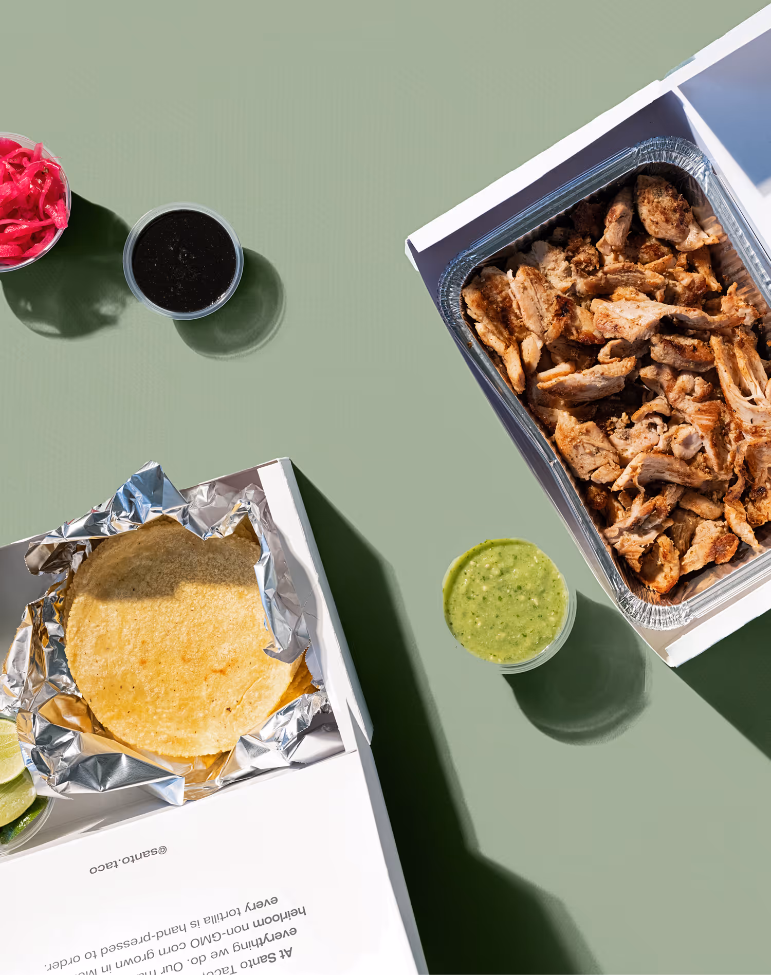 Three tacos on plates with grilled zucchini, meat topped with green salsa and lime, and diced vegetables, alongside a glass of iced yellow drink.