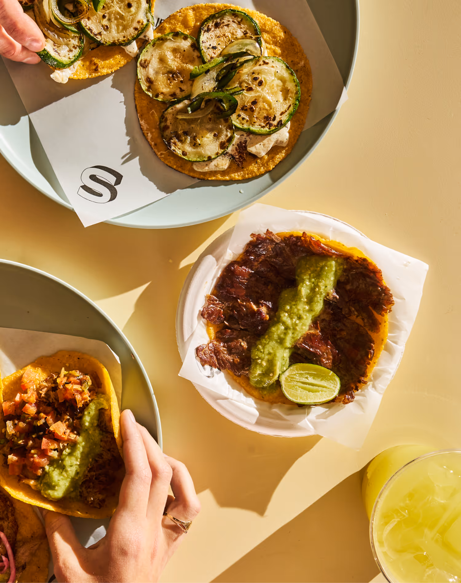 Three tacos on plates with grilled zucchini, meat topped with green salsa and lime, and diced vegetables, alongside a glass of iced yellow drink.