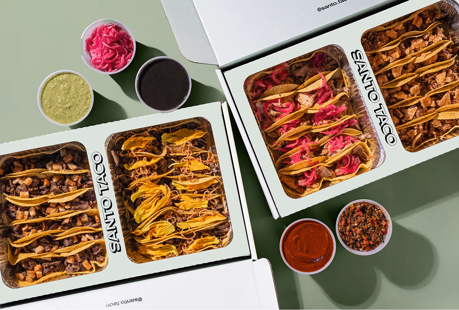 Person holding a taco with pickled onions and lime wedge above foil trays filled with tacos topped with green sauce and lime wedges.