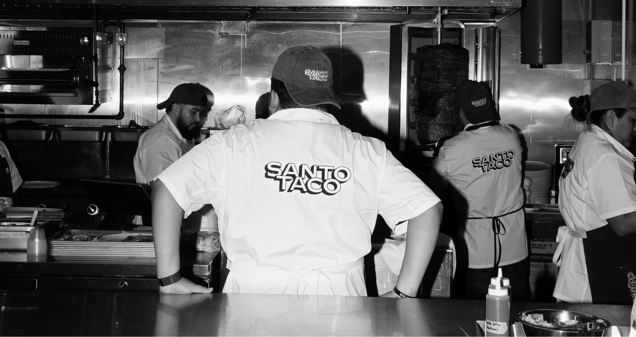 Santo Taco staff preparing food at Santo Taco restaurant, visible from behind with branded shirts and hats.