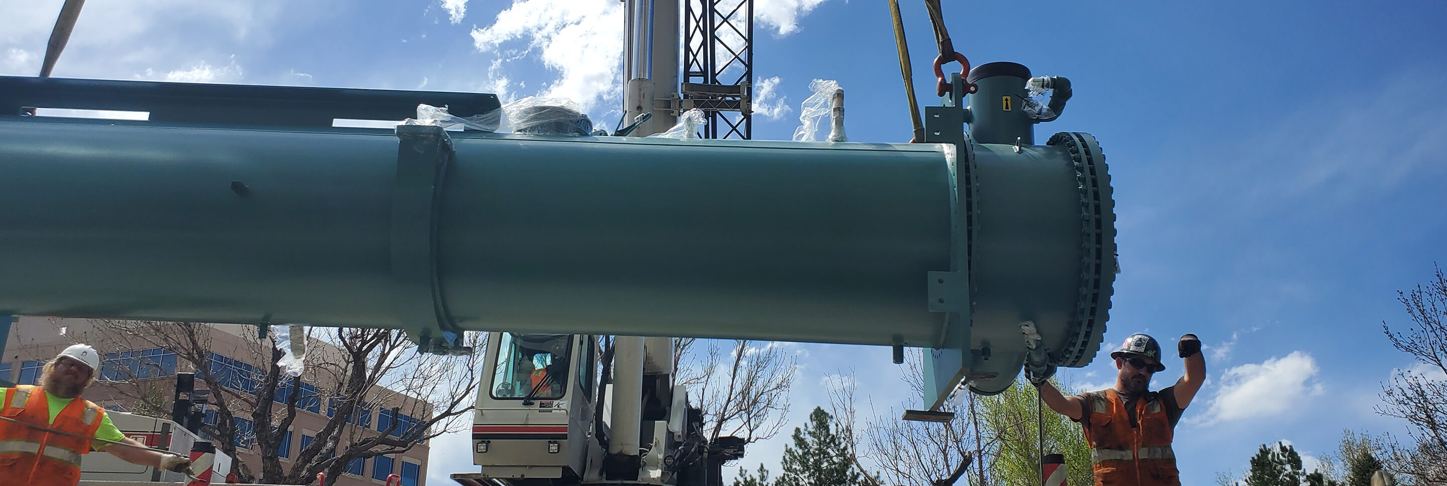 Two construction workers in safety gear guiding a large cylindrical industrial component being lifted by a crane outdoors.