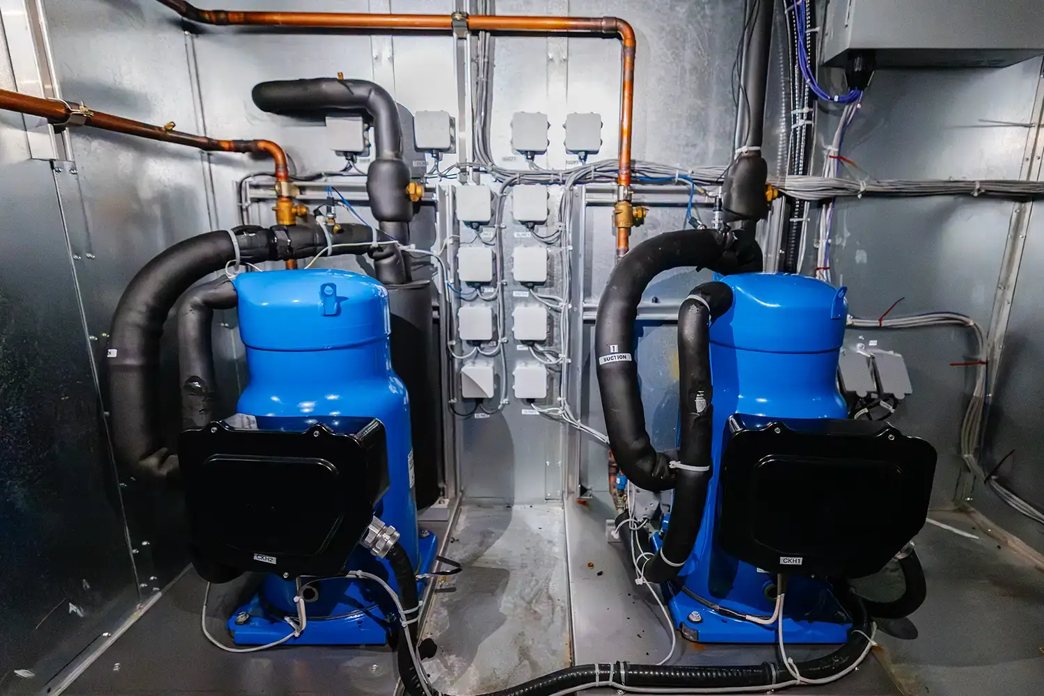Two blue industrial air conditioning compressors with insulated black pipes in a metallic equipment room.