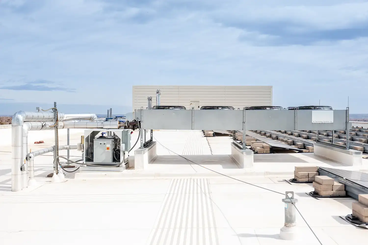 Industrial HVAC equipment including ductwork and fans installed on a flat white rooftop under a cloudy sky.