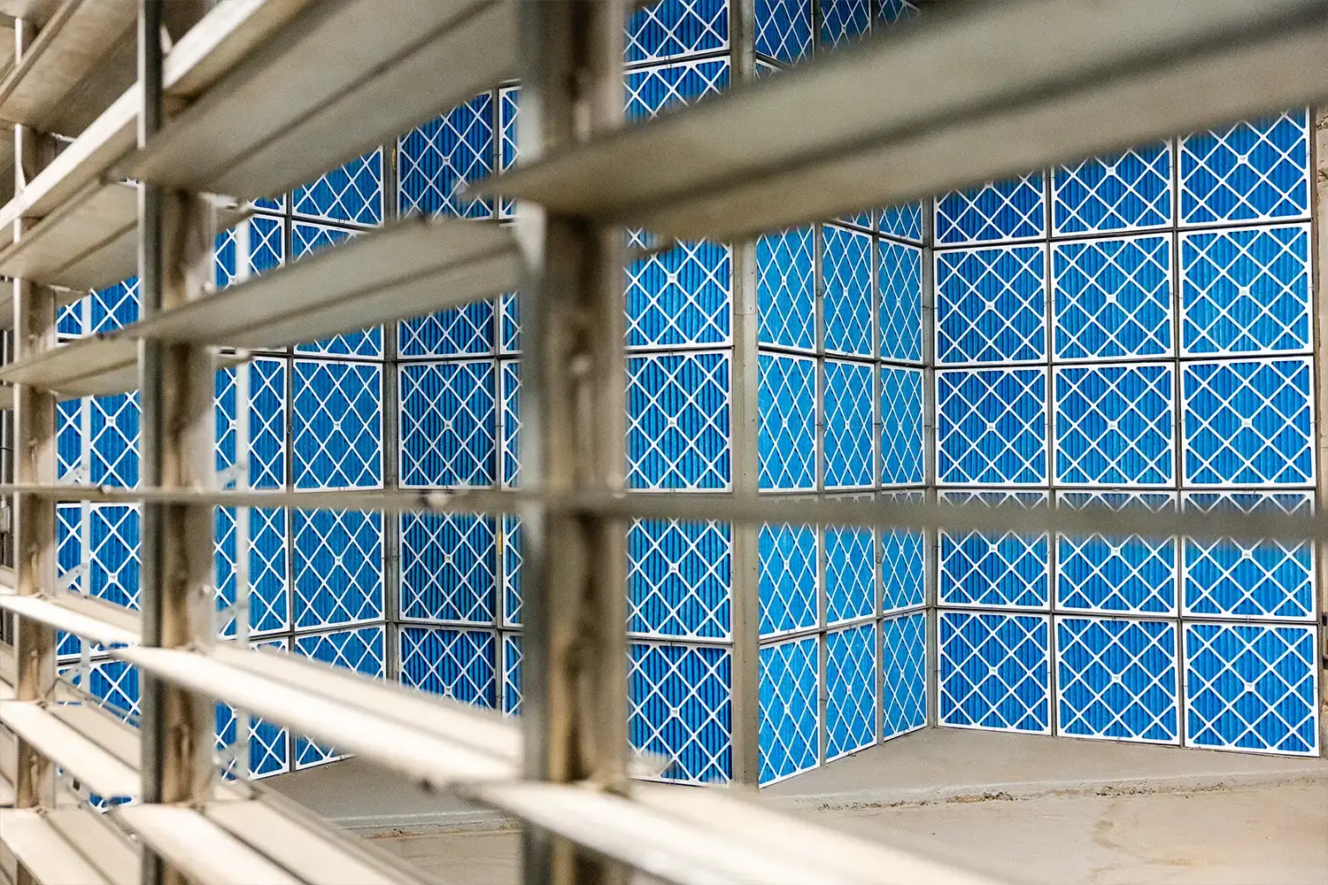 Blue air filters with white grid pattern inside a maintenance area, viewed through metal ventilation louver slats.