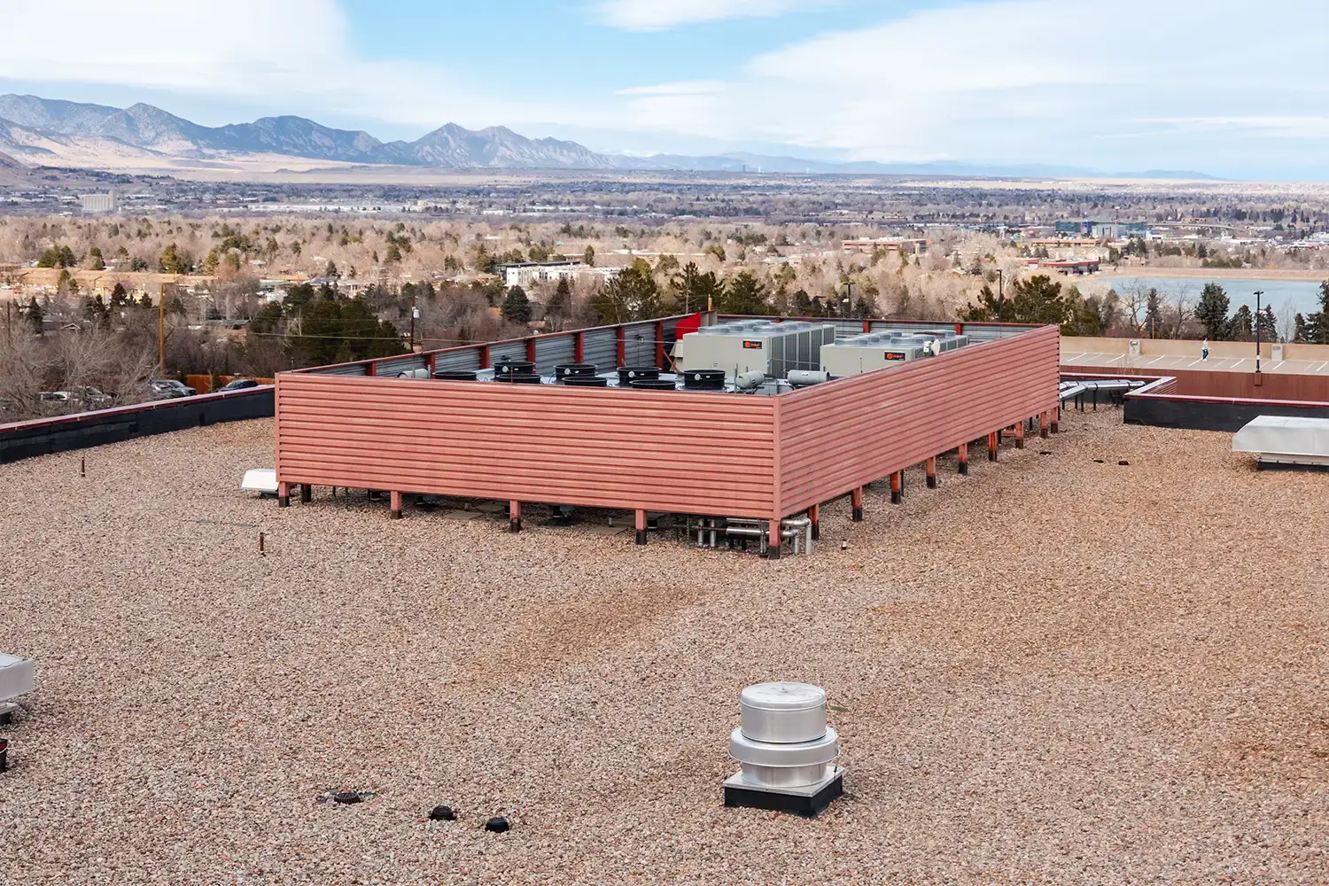 Rooftop HVAC units enclosed by a wooden fence on a gravel-covered building roof with a city and mountain landscape in the background.
