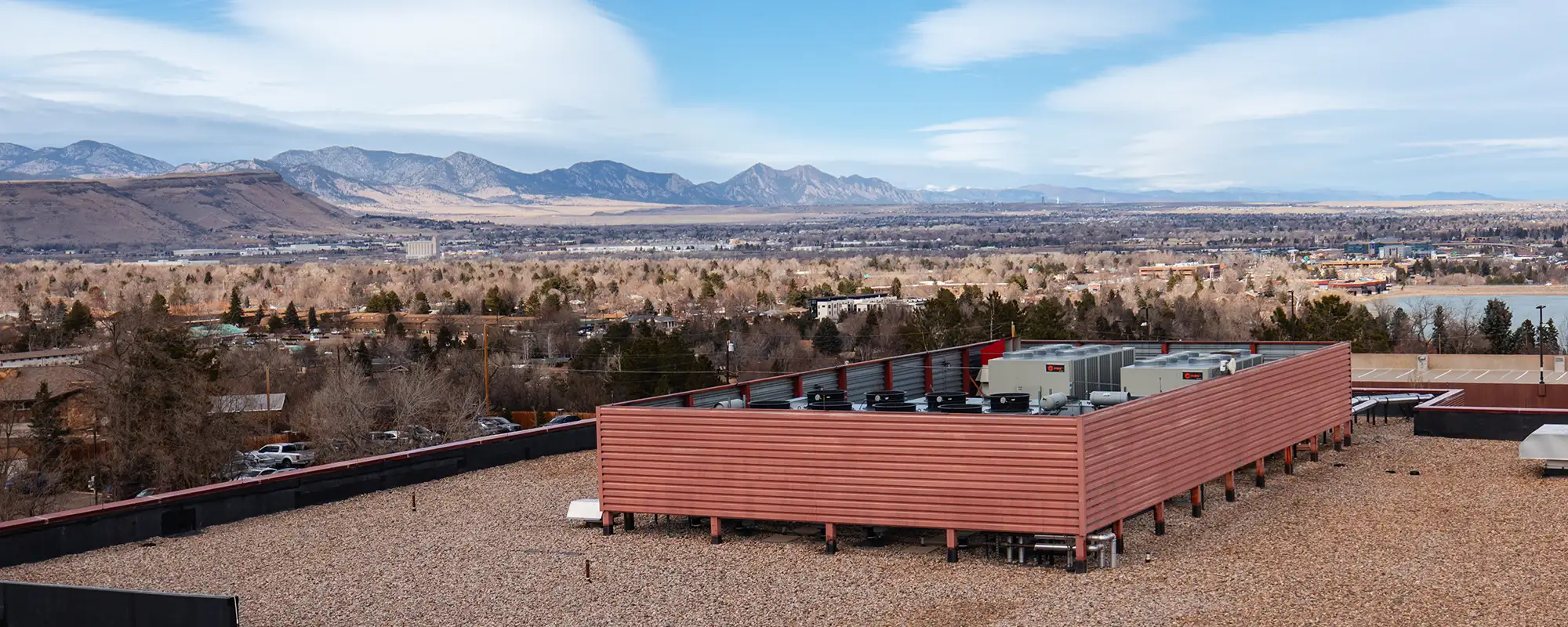 Urban landscape with a pebbled rooftop foreground featuring HVAC units and a backdrop of a suburban area with bare trees and distant mountain range.
