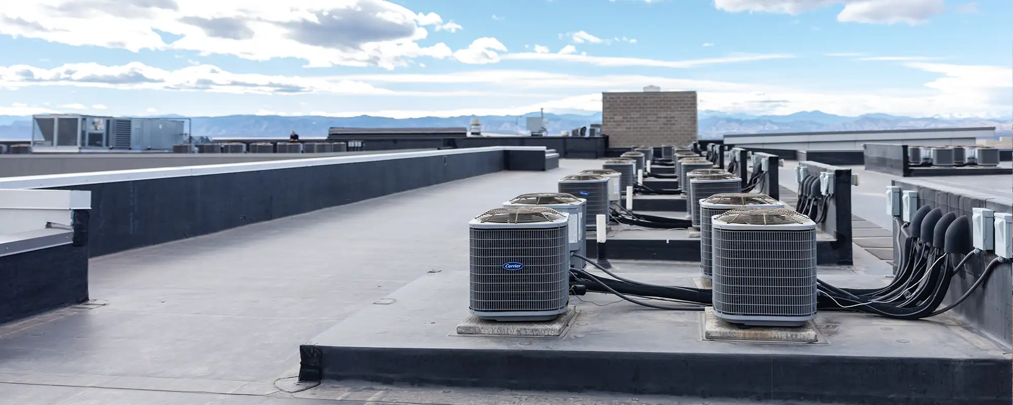 Row of Carrier air conditioning units installed on a flat rooftop with a mountain range in the background under a partly cloudy sky.