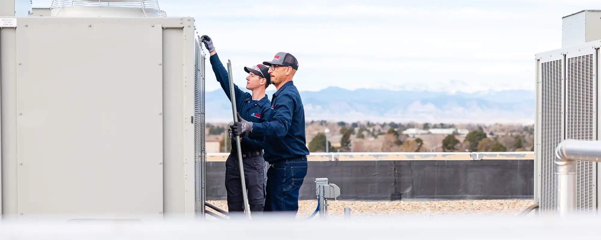Two technicians in navy uniforms and caps working on HVAC units on a rooftop with mountains in the background.