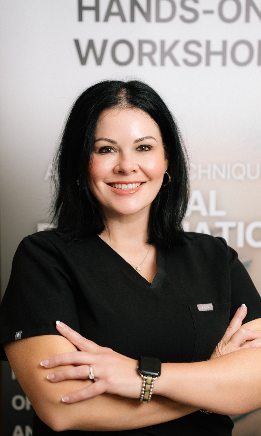 Middle-aged woman with shoulder-length blond hair wearing a black jacket and pearl necklace, smiling in front of a facial rejuvenation poster.