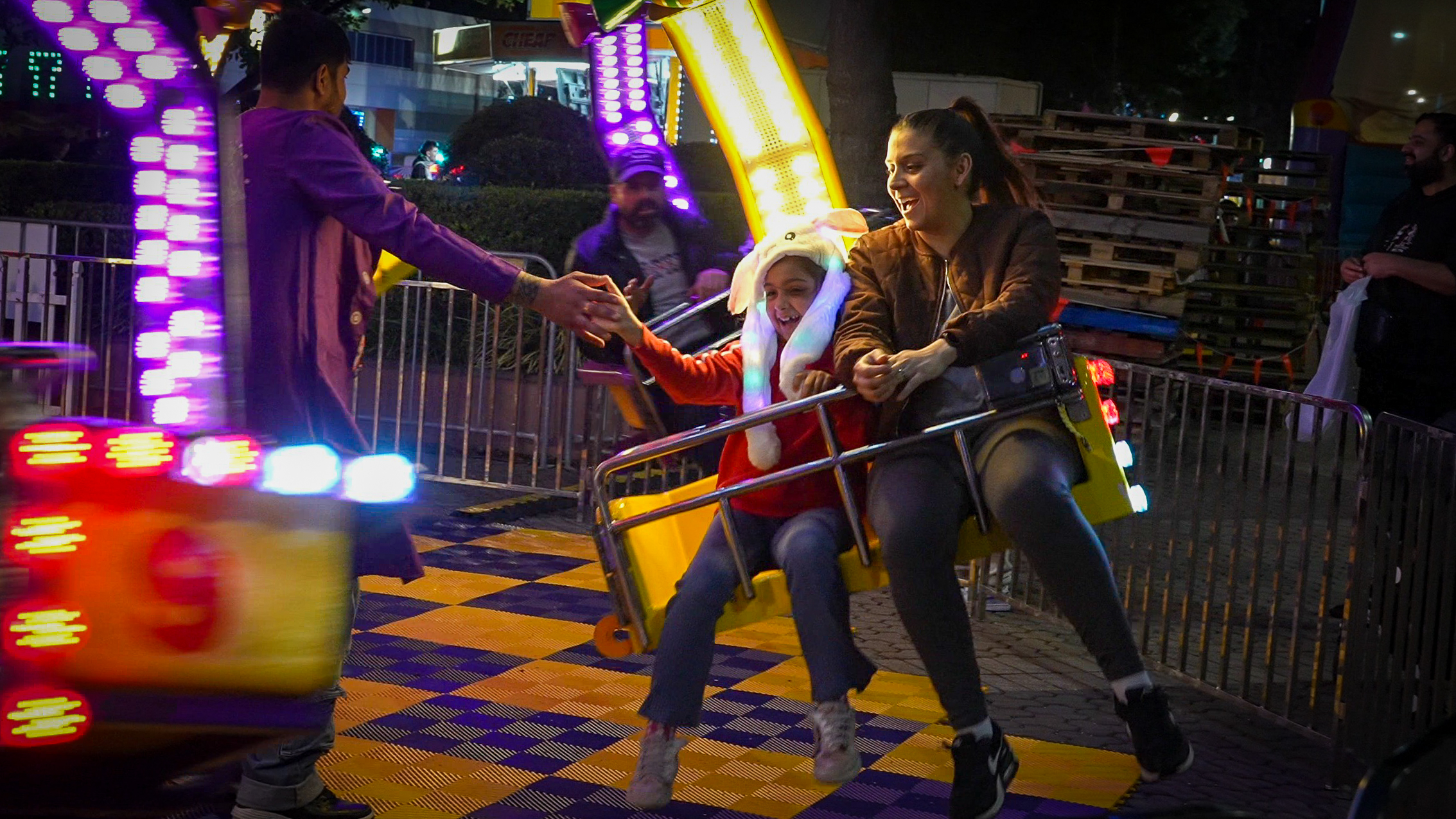 Child wearing a glowing bunny hat and an adult woman smiling on a yellow amusement ride seat at night, reaching for a man’s hand.