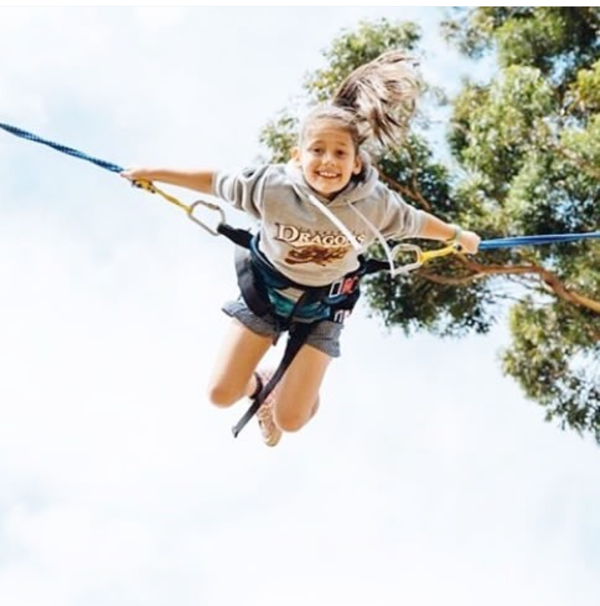 Smiling girl in a harness bouncing on a bungee trampoline with trees and sky in the background.