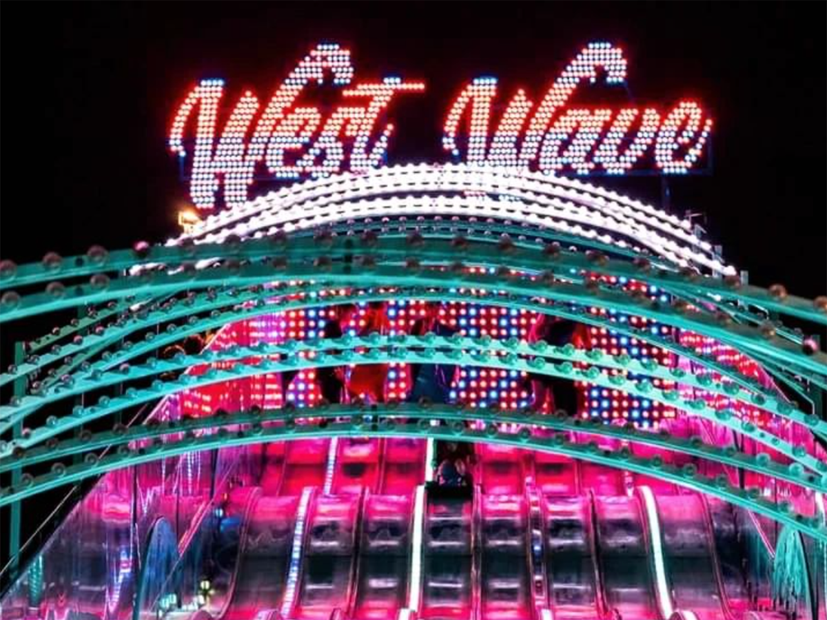 Illuminated West Wave amusement ride at night with neon lights and metal slide lanes.