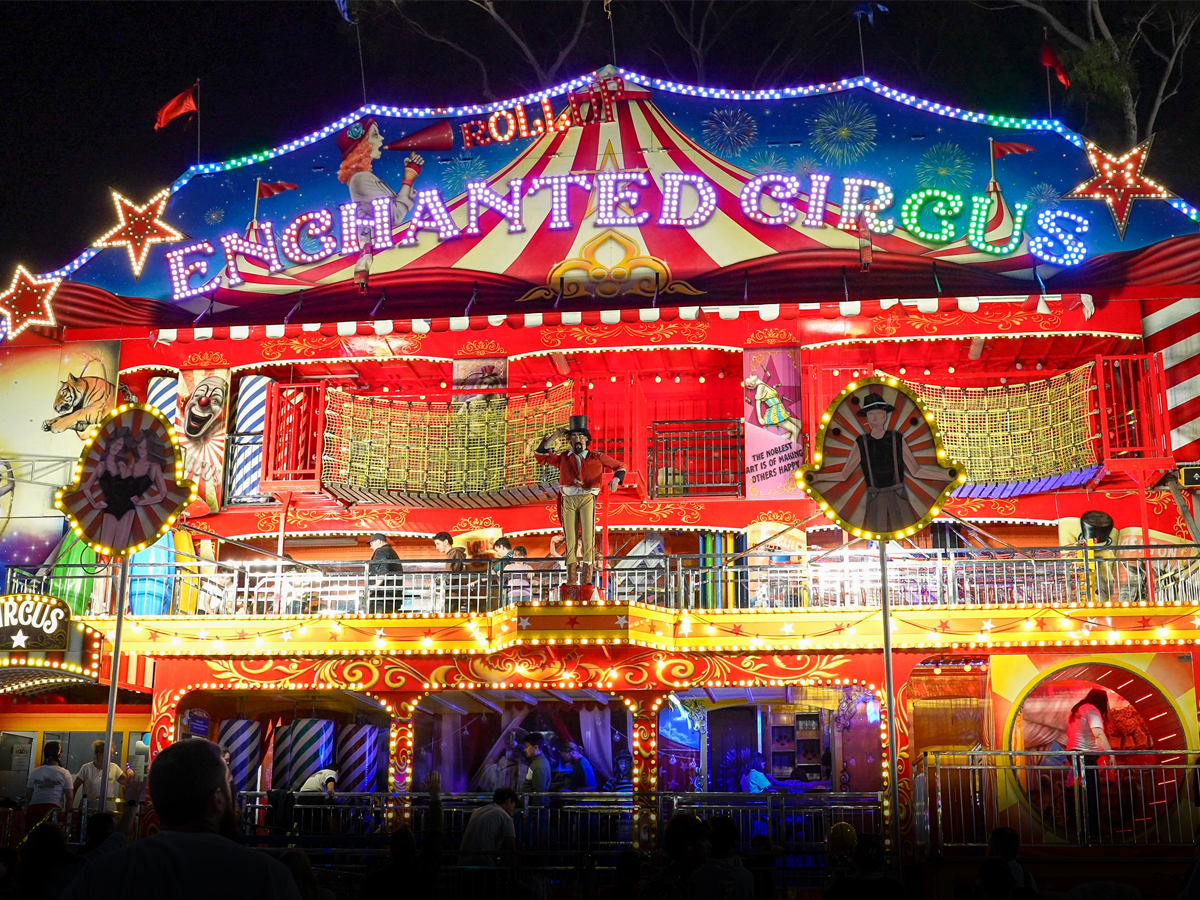 Brightly lit Enchanted Circus carnival facade at night with colorful lights, decorations, and people waiting.