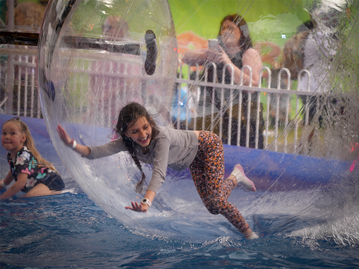 Young girl laughing inside a transparent inflatable water walking ball on a pool with another child nearby.
