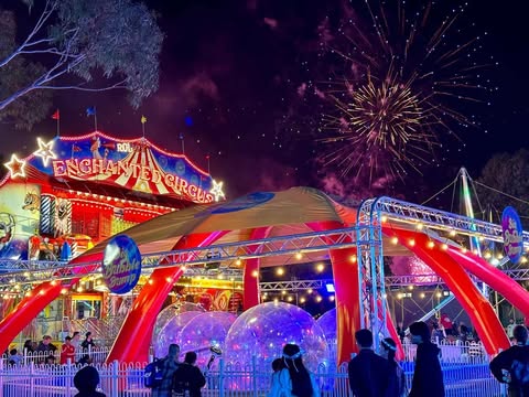 Night scene at a carnival with people watching large transparent spheres under a tent and fireworks exploding in the sky above a brightly lit Enchanted Circus sign.