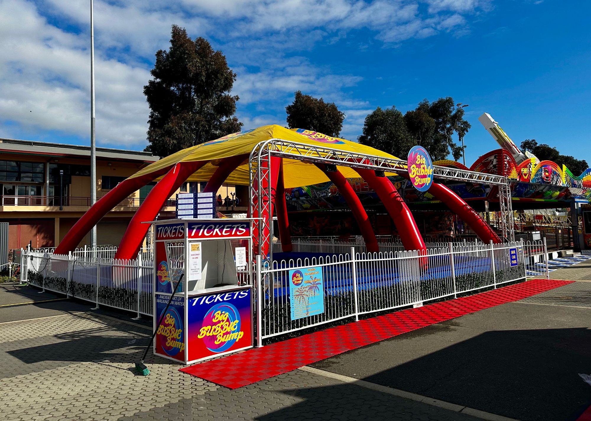 Outdoor amusement ride named Big Bubble Bump with a yellow and red inflatable structure and a ticket booth enclosed by a white metal fence.