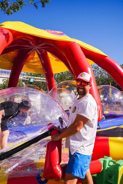 Man in white t-shirt, blue shorts, and cap smiling while holding an inflatable bubble structure at an outdoor event on a sunny day.