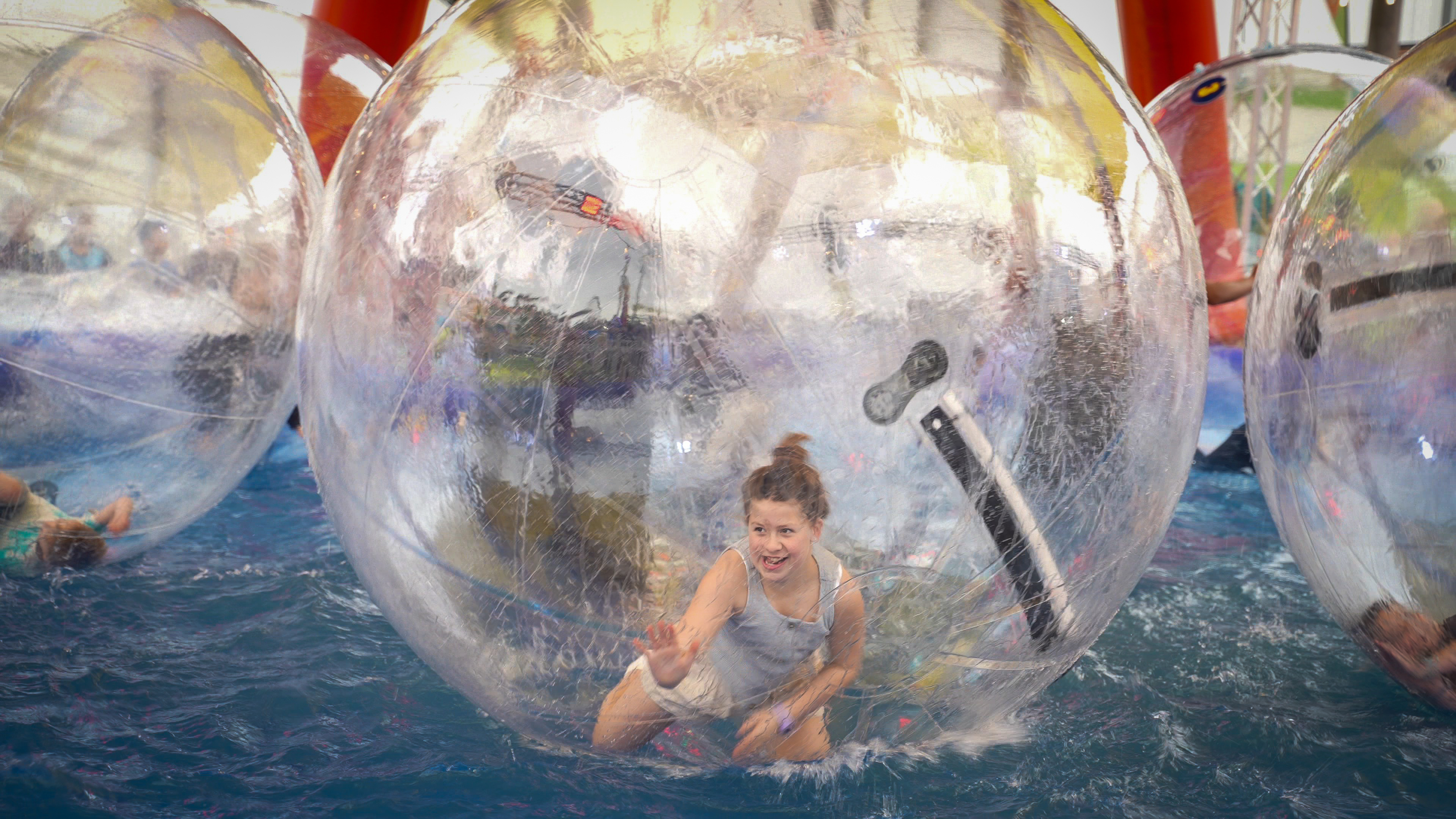 Girl inside a large transparent inflatable ball floating on water, smiling and reaching forward.