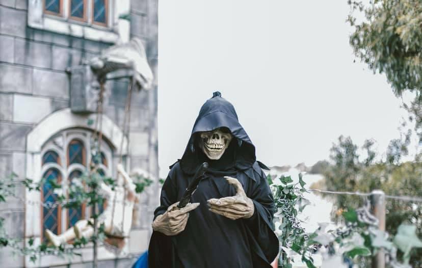 Person dressed as a grim reaper with a skeletal mask and black hooded robe holding a scythe, standing outdoors near a stone building and greenery.