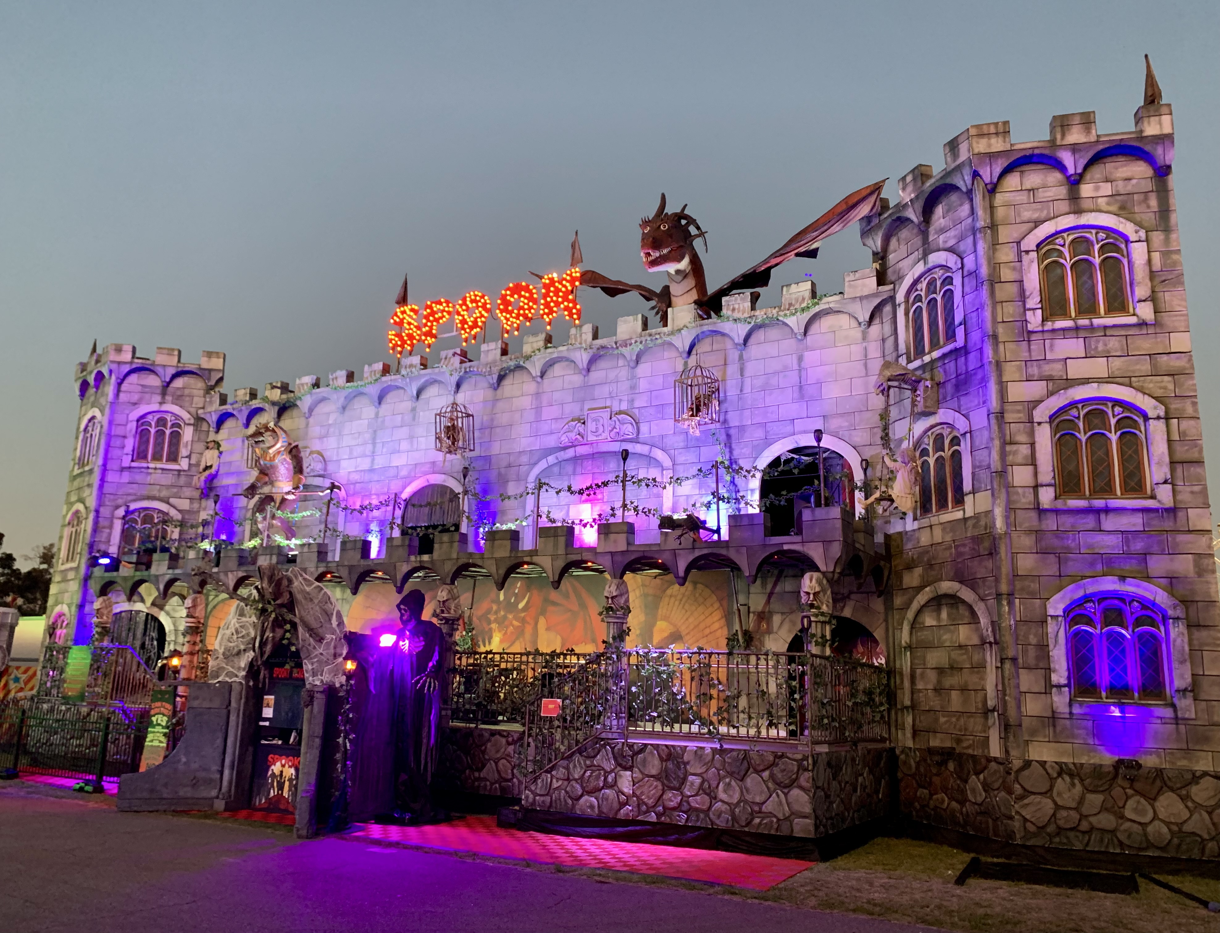 Illuminated haunted castle facade at dusk decorated with a dragon figure, skeletons in cages, and a glowing red 'SPOOK' sign.