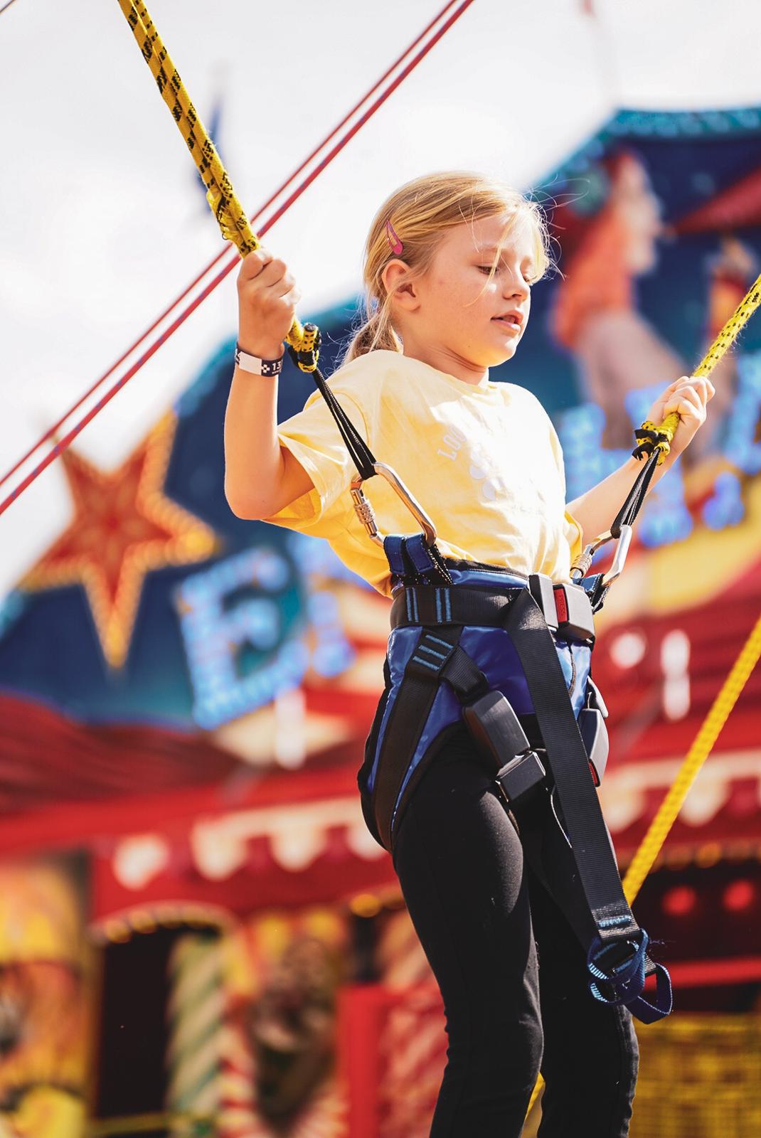 Young girl in a safety harness holding yellow bungee cords at a carnival or amusement park.