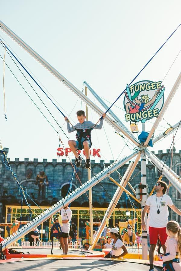 Boy jumping on a bungee trampoline at an outdoor amusement park with staff supervising.