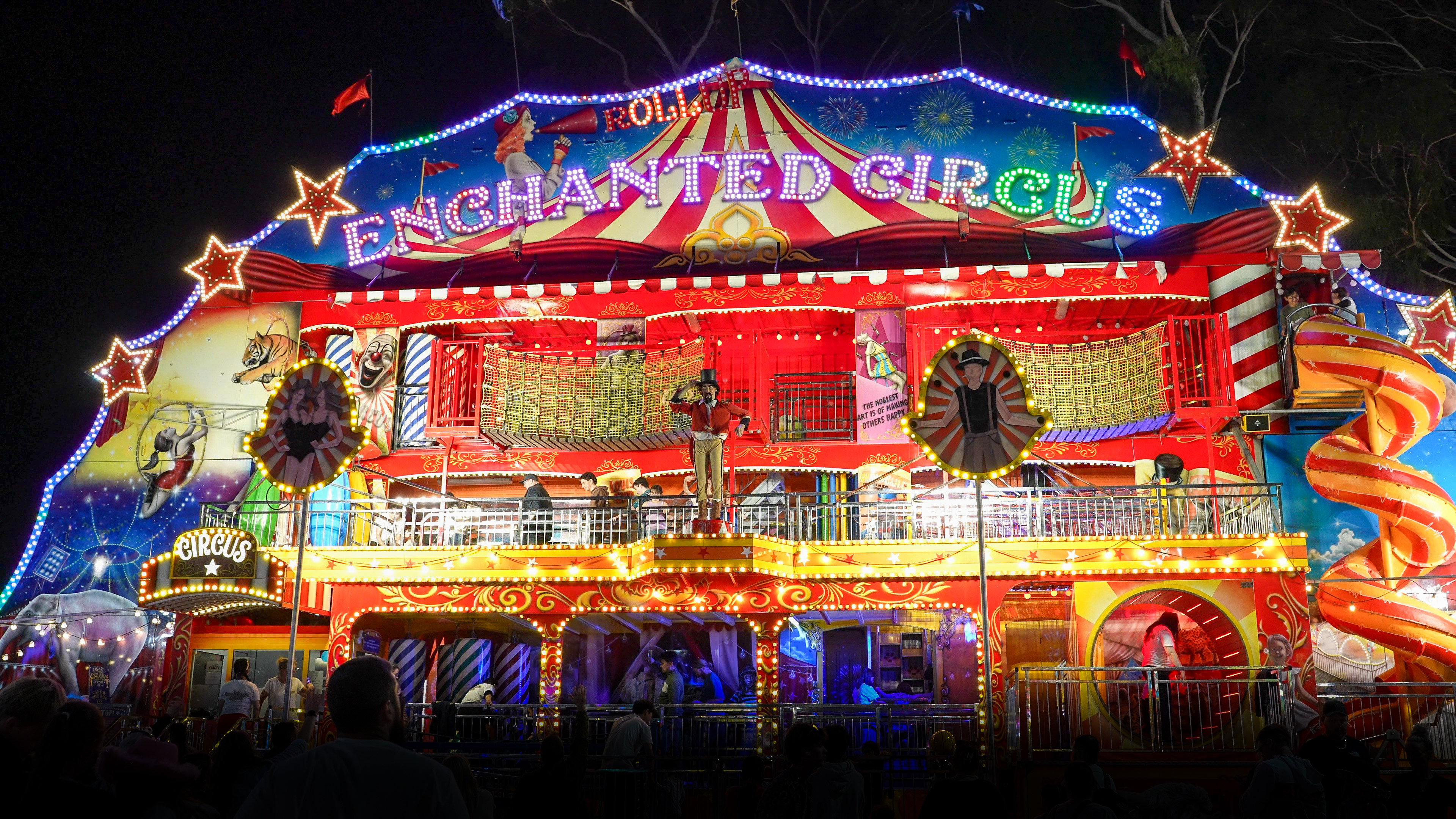 Brightly lit two-story carnival funhouse at night with 'Enchanted Circus' sign, various circus-themed decorations, and a large orange spiral slide.