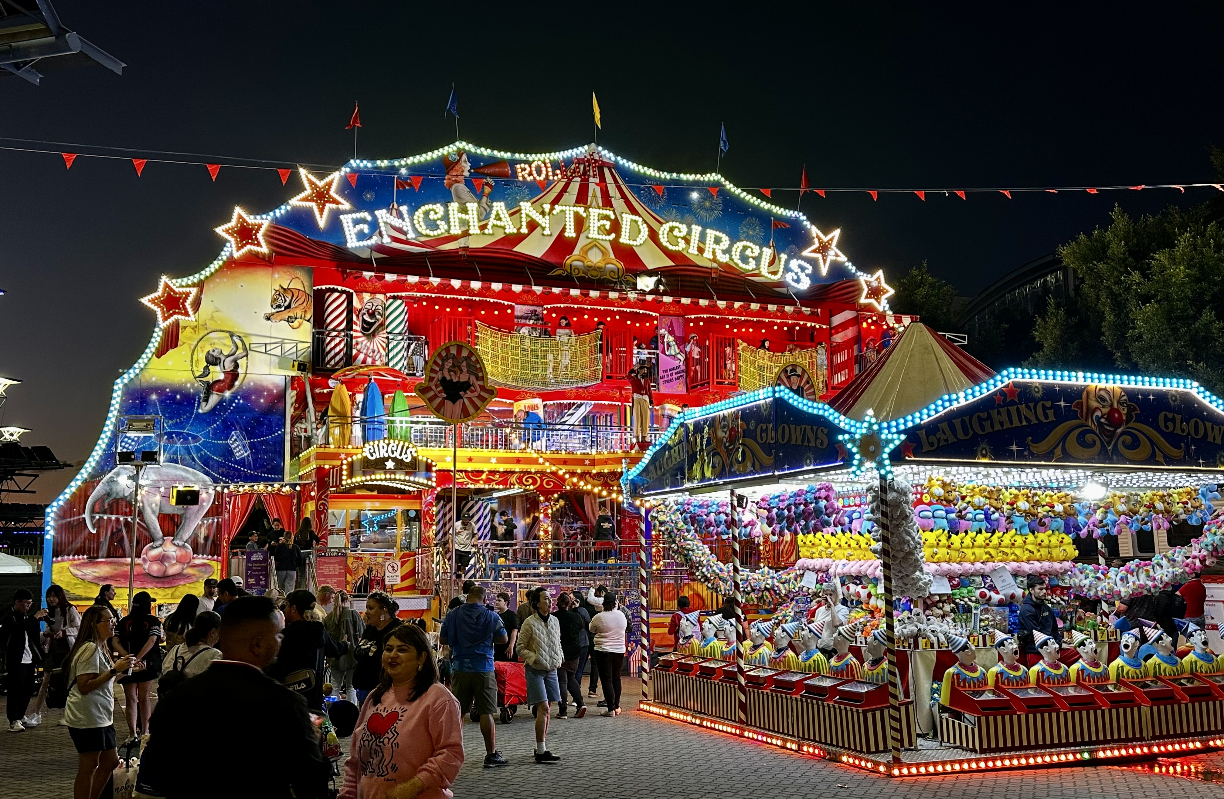 Night scene at a brightly lit circus carnival with an Enchanted Circus building and a Laughing Clown game booth with stuffed animal prizes.