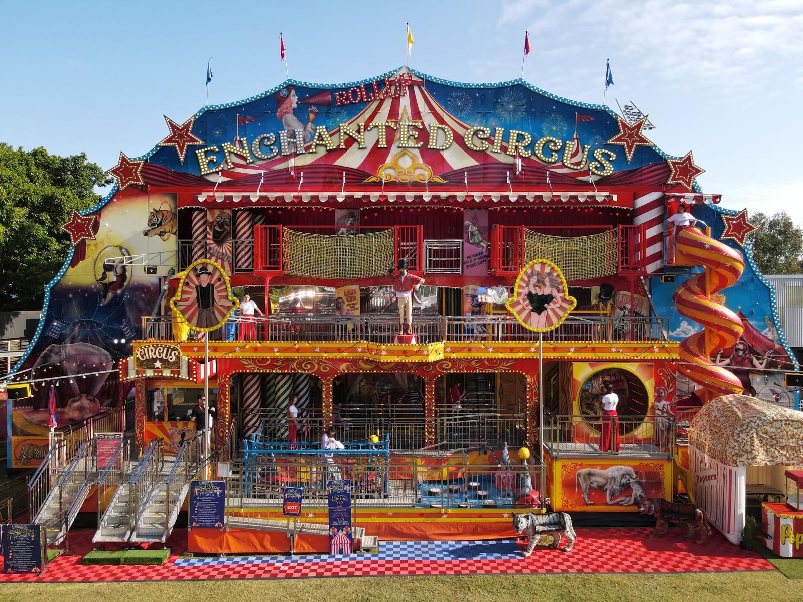 Colorful multi-level amusement funhouse named Enchanted Circus with slides, climbing nets, and circus-themed decorations.