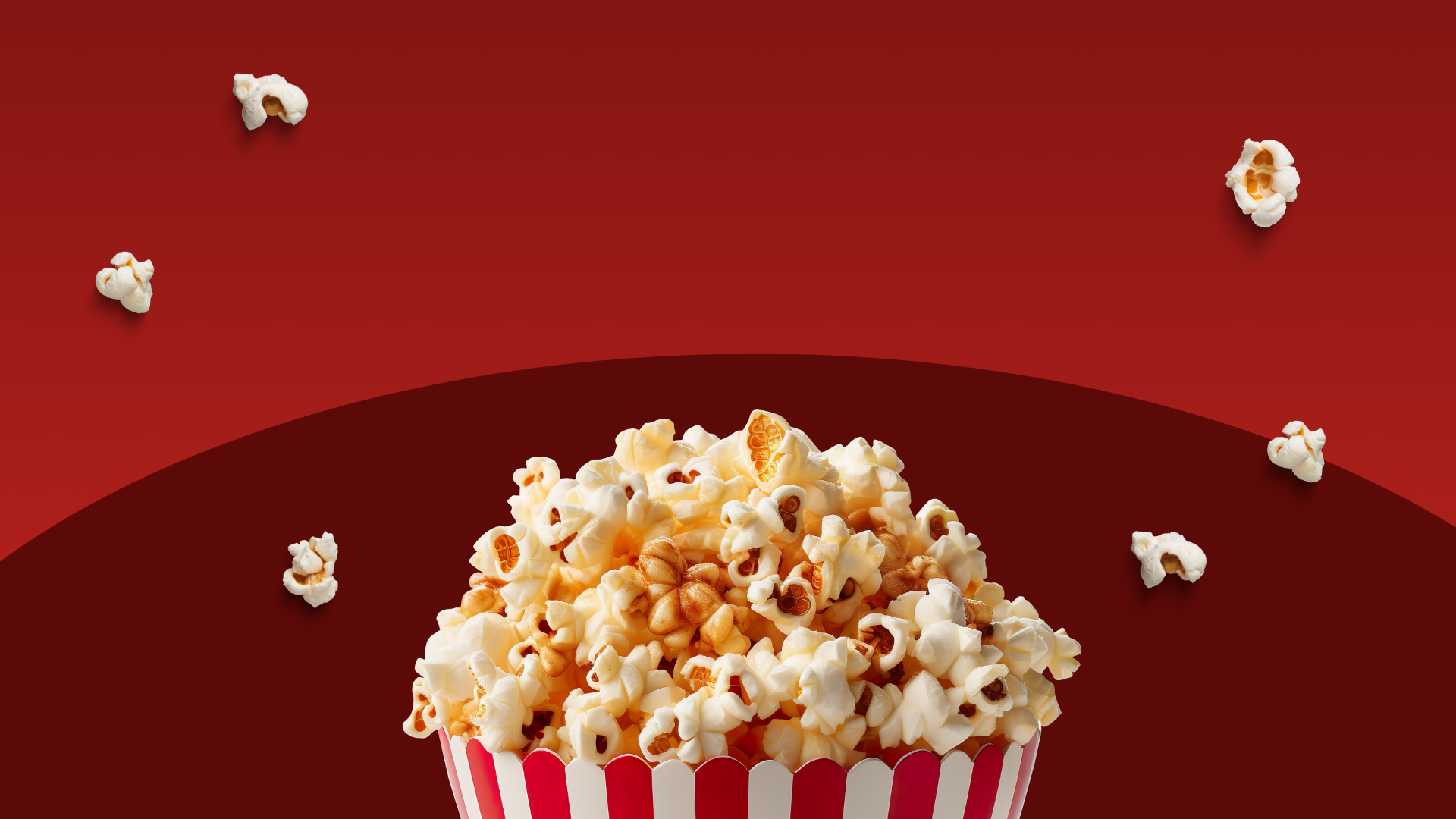 Close-up of a red and white striped container filled with buttered popcorn against a red background with scattered popcorn pieces.