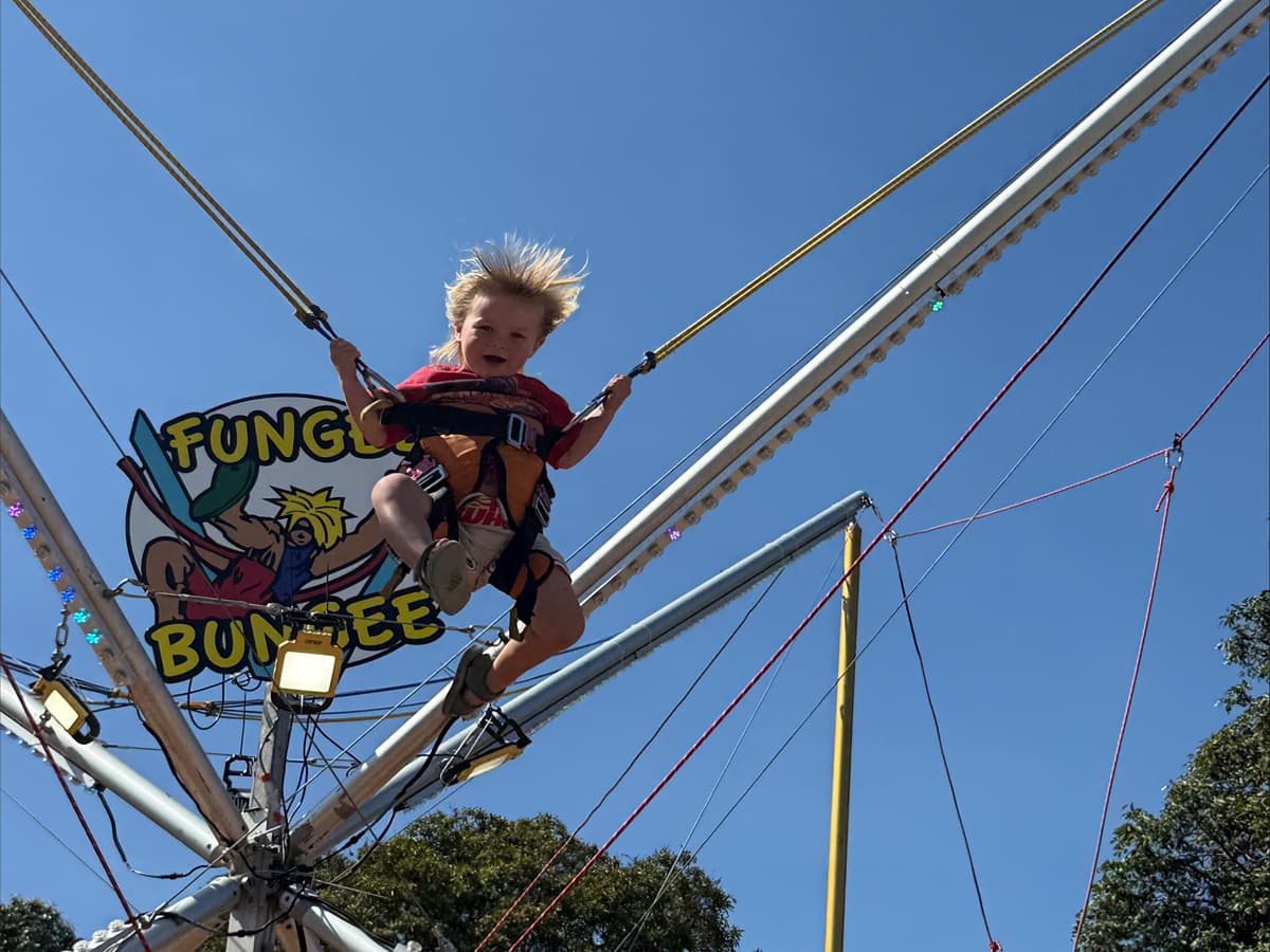 Child with windswept hair bouncing on a bungee trampoline against a clear blue sky.