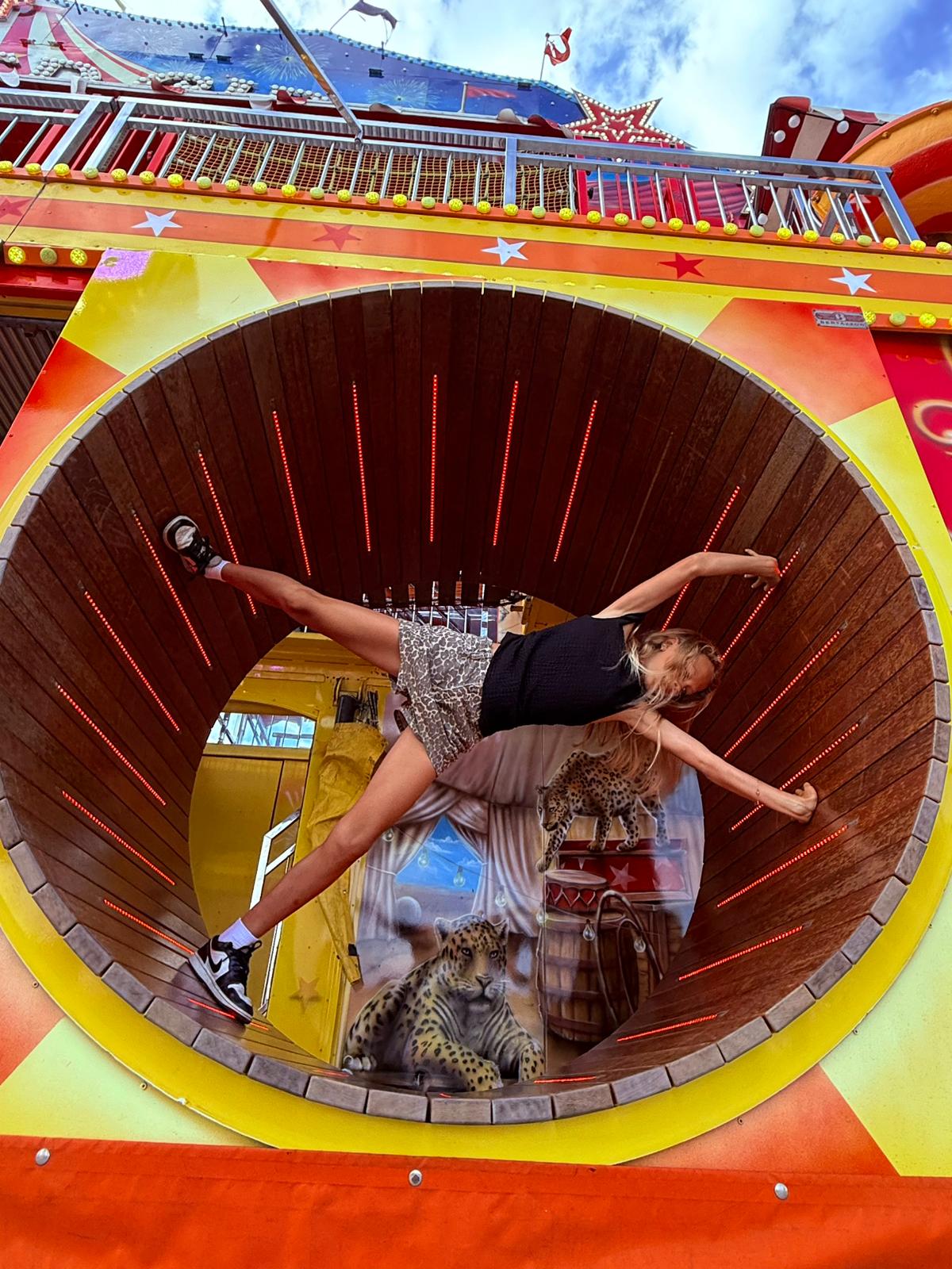 Girl balancing horizontally inside a large vertical wooden wheel decorated with leopard paintings and colorful carnival elements.