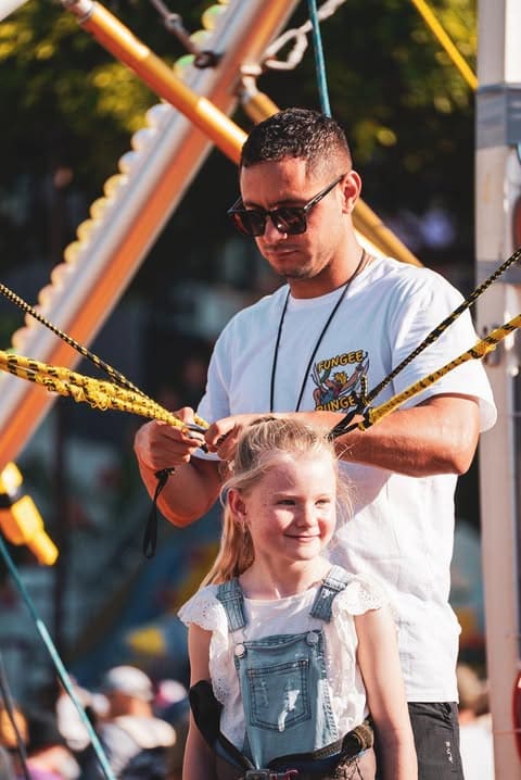Man securing harness for young girl at a bungee trampoline setup outdoors.