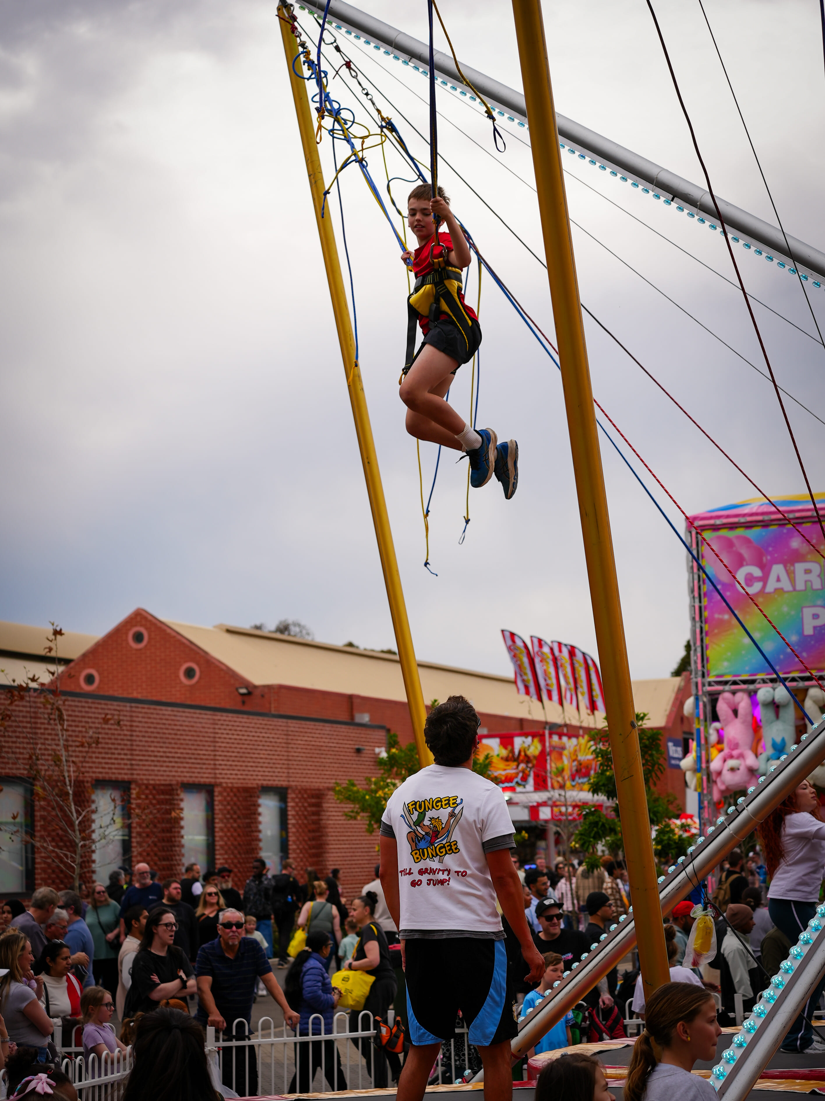 Boy suspended mid-air on bungee trampoline with an instructor watching below at a crowded outdoor fair.