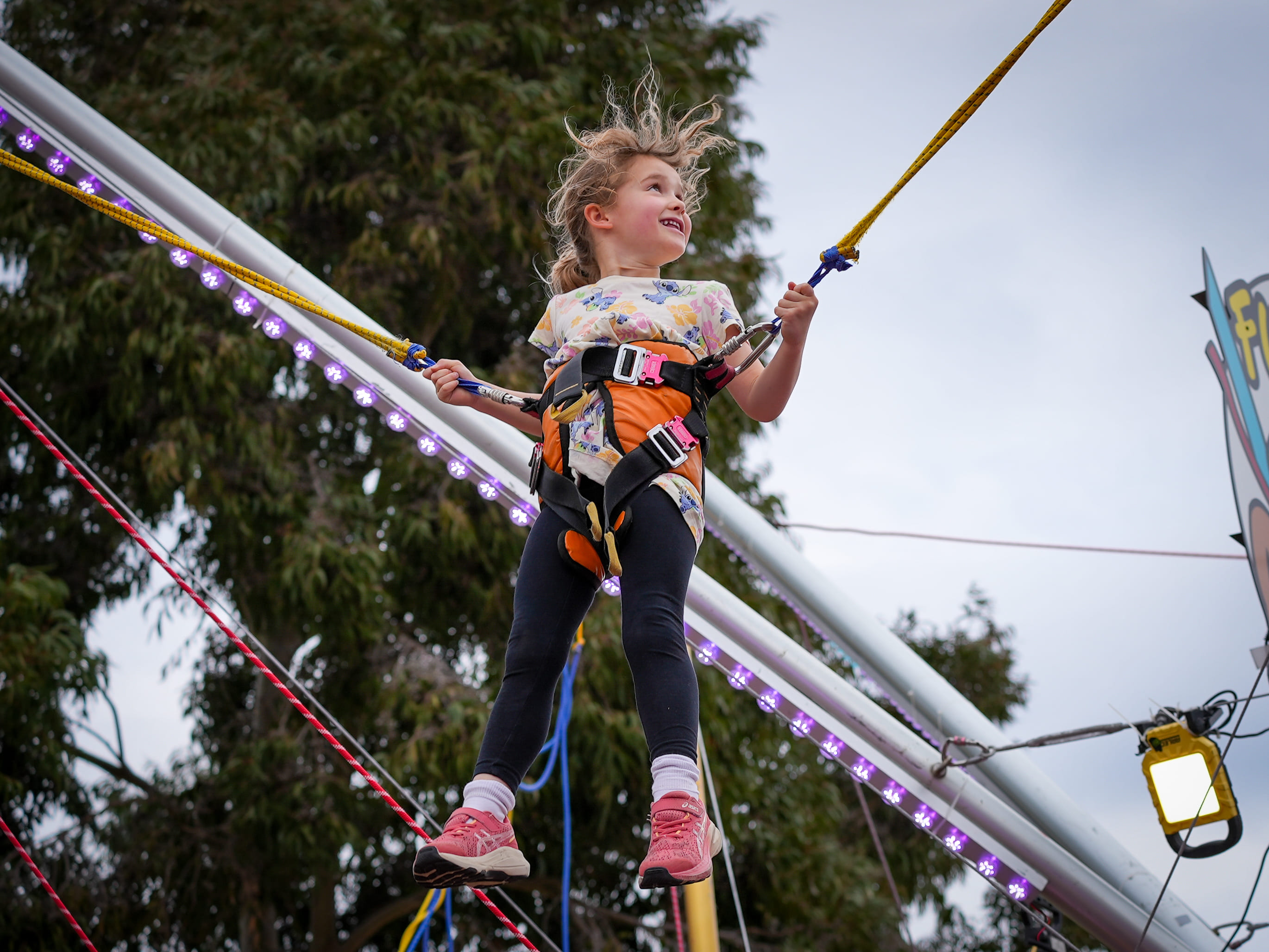 A young girl wearing a safety harness bounces on a trampoline bungee ride at an outdoor fair.