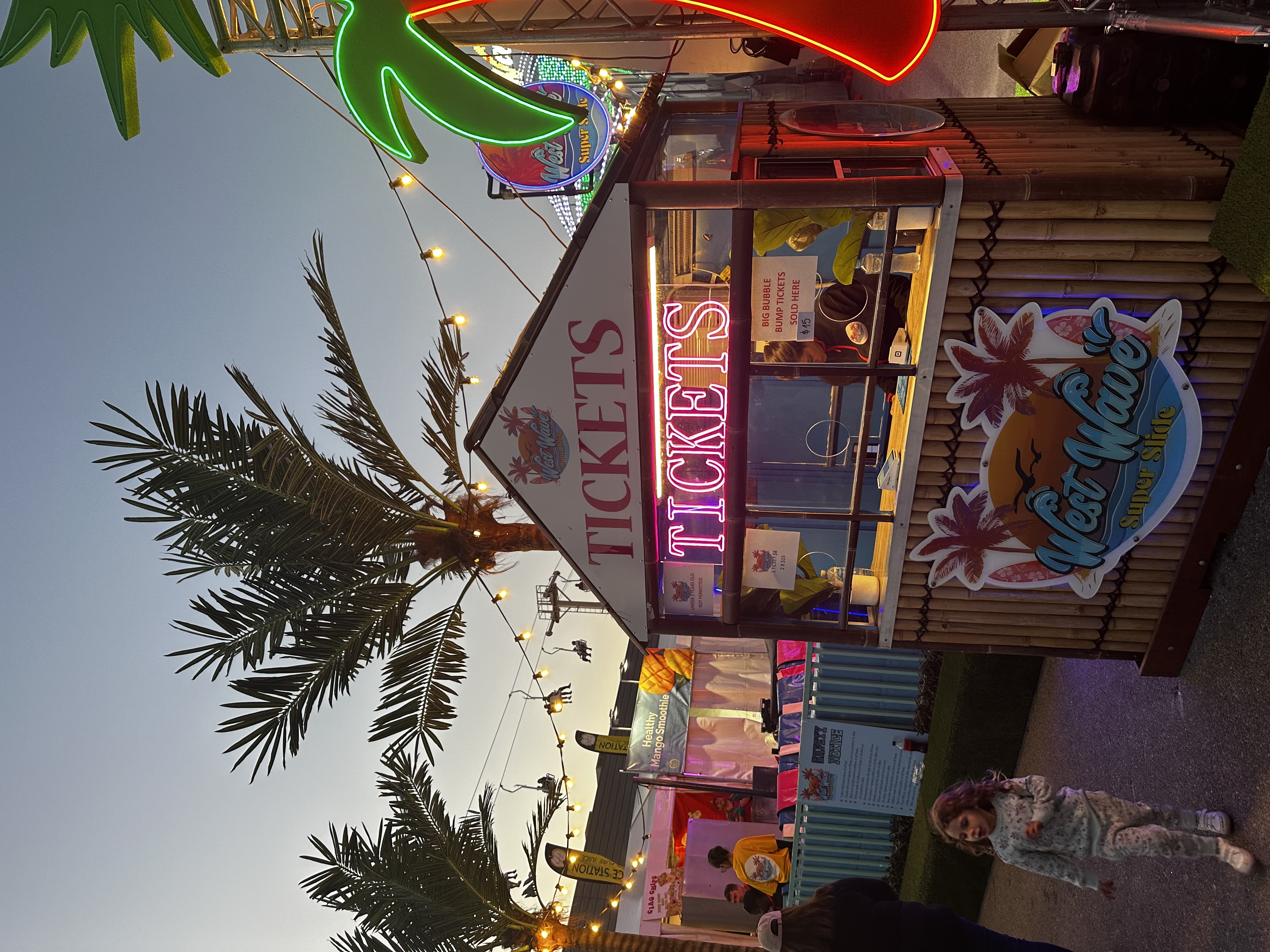 Ticket booth with neon 'TICKETS' sign at West Wave Super Slide attraction, with palm trees, string lights, and a child walking nearby.